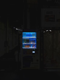 A vending machine is positioned in a dimly lit urban setting, surrounded by shadows. The machine is brightly illuminated, highlighting its colorful array of beverages behind the glass display. The contrast between the light of the machine and the surrounding darkness is stark.
