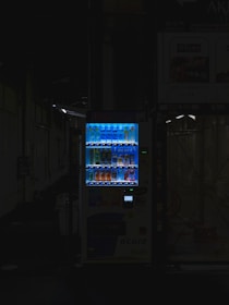 A vending machine is positioned in a dimly lit urban setting, surrounded by shadows. The machine is brightly illuminated, highlighting its colorful array of beverages behind the glass display. The contrast between the light of the machine and the surrounding darkness is stark.