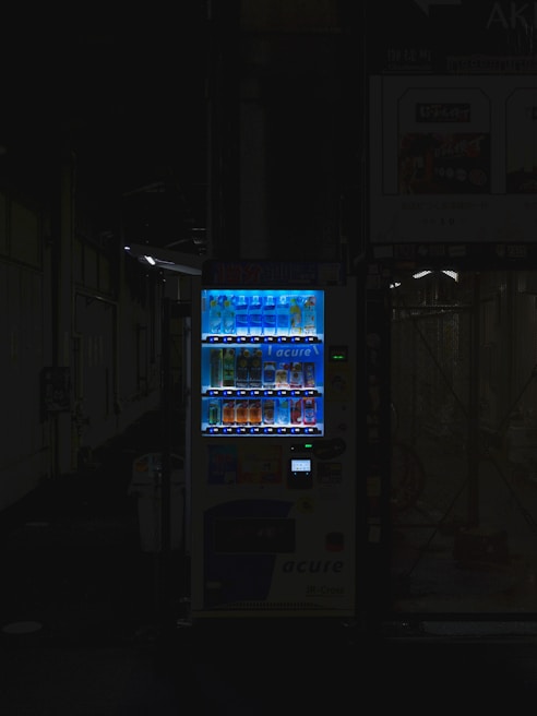 A vending machine is positioned in a dimly lit urban setting, surrounded by shadows. The machine is brightly illuminated, highlighting its colorful array of beverages behind the glass display. The contrast between the light of the machine and the surrounding darkness is stark.