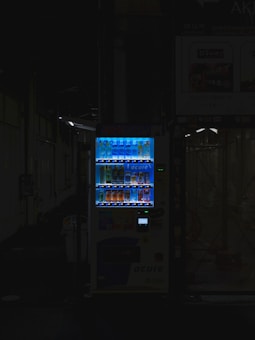 A vending machine is positioned in a dimly lit urban setting, surrounded by shadows. The machine is brightly illuminated, highlighting its colorful array of beverages behind the glass display. The contrast between the light of the machine and the surrounding darkness is stark.