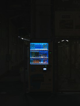 A vending machine is positioned in a dimly lit urban setting, surrounded by shadows. The machine is brightly illuminated, highlighting its colorful array of beverages behind the glass display. The contrast between the light of the machine and the surrounding darkness is stark.