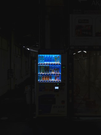 A vending machine is positioned in a dimly lit urban setting, surrounded by shadows. The machine is brightly illuminated, highlighting its colorful array of beverages behind the glass display. The contrast between the light of the machine and the surrounding darkness is stark.