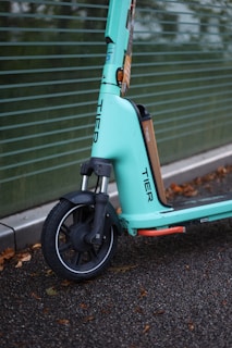A close-up view of an electric scooter with a teal color scheme parked on a wet asphalt surface. The scooter is branded with the word 'TIER' on its front panel, and the image captures the front wheel and part of the handlebar area. Fallen leaves are scattered on the ground, and a green, horizontal-lined backdrop is partially visible.
