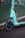 A close-up view of an electric scooter with a teal color scheme parked on a wet asphalt surface. The scooter is branded with the word 'TIER' on its front panel, and the image captures the front wheel and part of the handlebar area. Fallen leaves are scattered on the ground, and a green, horizontal-lined backdrop is partially visible.