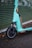 A close-up view of an electric scooter with a teal color scheme parked on a wet asphalt surface. The scooter is branded with the word 'TIER' on its front panel, and the image captures the front wheel and part of the handlebar area. Fallen leaves are scattered on the ground, and a green, horizontal-lined backdrop is partially visible.
