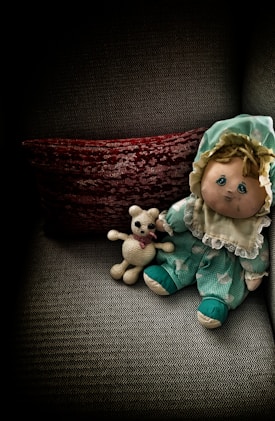 A plush doll dressed in a green outfit with a bonnet sits on a textured gray armchair beside a small crocheted teddy bear. Behind them is a deep red textured pillow, adding contrast to the scene with a cozy and nostalgic feel.