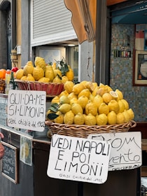 A street vendor display showcasing baskets overflowing with large, bright yellow lemons. Handwritten signs in Italian are prominently visible, advertising the lemons with playful phrases. The scene is set against a backdrop of a closed storefront with a striped awning and tiled wall.