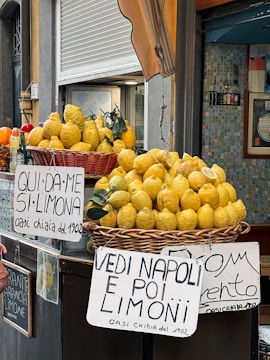 A street vendor display showcasing baskets overflowing with large, bright yellow lemons. Handwritten signs in Italian are prominently visible, advertising the lemons with playful phrases. The scene is set against a backdrop of a closed storefront with a striped awning and tiled wall.
