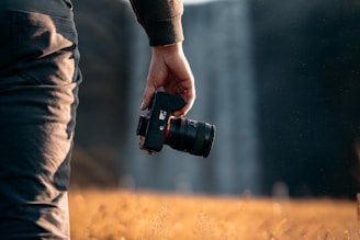 A person holds a camera in their right hand, standing in a grassy field with a blurred waterfall in the background. The lighting creates a warm, golden glow on the scene, emphasizing the natural setting.
