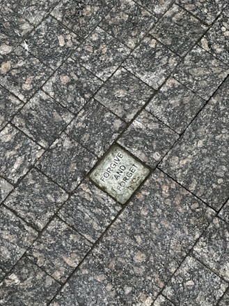 A stone tile pavement with a singular stone in the center engraved with the words 'FORGIVE AND FORGET.' The surrounding tiles are textured with a mix of gray and dark tones, giving a rugged appearance.