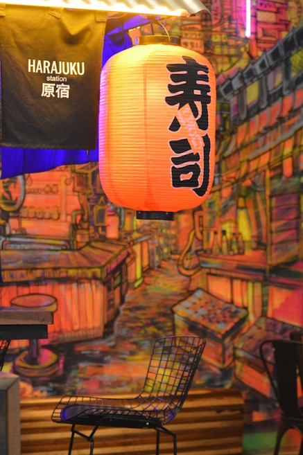 A vibrant setting with an orange Japanese lantern displaying black kanji characters, hanging above a scenic mural depicting an illuminated urban street. A black metal chair is positioned beneath the lantern, and a fabric banner labeled 'Harajuku station' is visible.