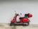 A bright red scooter with a sleek design is parked against a pale wall. The scooter features a black seat and a top case storage compartment at the rear. The overall setting is minimalistic, with a neutral background highlighting the vibrant color of the scooter.
