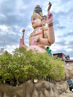A large, intricately detailed statue depicted in a seated position, resembling a deity with multiple arms, holding various symbolic items. The statue is adorned with an ornate headdress and jewelry, surrounded by greenery and artificial rock formations. The background features a cloudy sky and nearby structures.