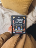 A hand with red-painted nails is holding a book titled 'My Hygge Home: How to Make Home Your Happy Place' by Meik Wiking. The person is dressed in a beige sweater and tan coat. The background consists of a fluffy beige blanket and a textured yellow pillow.