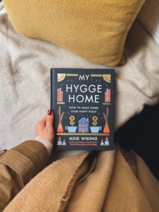 A hand with red-painted nails is holding a book titled 'My Hygge Home: How to Make Home Your Happy Place' by Meik Wiking. The person is dressed in a beige sweater and tan coat. The background consists of a fluffy beige blanket and a textured yellow pillow.