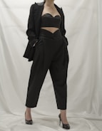 A person wearing a stylish black outfit, comprising a tailored blazer, a cutout top, and high-waisted pants. The ensemble is complemented by black high-heeled shoes. The background is a neutral white, which emphasizes the dark clothing.