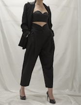 A person wearing a stylish black outfit, comprising a tailored blazer, a cutout top, and high-waisted pants. The ensemble is complemented by black high-heeled shoes. The background is a neutral white, which emphasizes the dark clothing.
