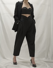 A person wearing a stylish black outfit, comprising a tailored blazer, a cutout top, and high-waisted pants. The ensemble is complemented by black high-heeled shoes. The background is a neutral white, which emphasizes the dark clothing.