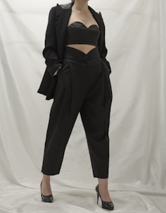 A person wearing a stylish black outfit, comprising a tailored blazer, a cutout top, and high-waisted pants. The ensemble is complemented by black high-heeled shoes. The background is a neutral white, which emphasizes the dark clothing.