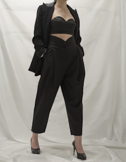 A person wearing a stylish black outfit, comprising a tailored blazer, a cutout top, and high-waisted pants. The ensemble is complemented by black high-heeled shoes. The background is a neutral white, which emphasizes the dark clothing.