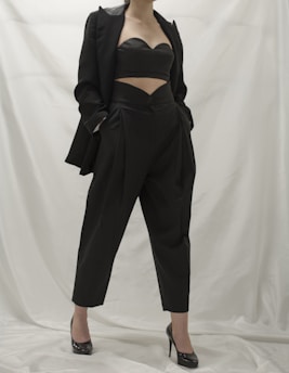 A person wearing a stylish black outfit, comprising a tailored blazer, a cutout top, and high-waisted pants. The ensemble is complemented by black high-heeled shoes. The background is a neutral white, which emphasizes the dark clothing.