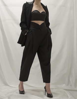A person wearing a stylish black outfit, comprising a tailored blazer, a cutout top, and high-waisted pants. The ensemble is complemented by black high-heeled shoes. The background is a neutral white, which emphasizes the dark clothing.