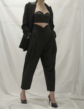 A person wearing a stylish black outfit, comprising a tailored blazer, a cutout top, and high-waisted pants. The ensemble is complemented by black high-heeled shoes. The background is a neutral white, which emphasizes the dark clothing.