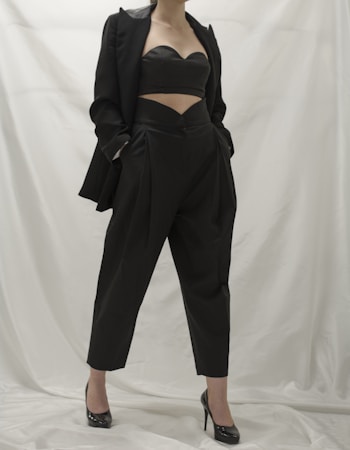 A person wearing a stylish black outfit, comprising a tailored blazer, a cutout top, and high-waisted pants. The ensemble is complemented by black high-heeled shoes. The background is a neutral white, which emphasizes the dark clothing.