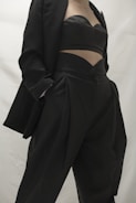 A person in a stylish black outfit, featuring a blazer and a uniquely designed top paired with high-waisted pants. The clothing appears sophisticated and tailored, emphasizing modern fashion trends.