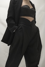 A person in a stylish black outfit, featuring a blazer and a uniquely designed top paired with high-waisted pants. The clothing appears sophisticated and tailored, emphasizing modern fashion trends.
