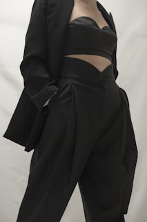 A person in a stylish black outfit, featuring a blazer and a uniquely designed top paired with high-waisted pants. The clothing appears sophisticated and tailored, emphasizing modern fashion trends.