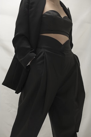A person in a stylish black outfit, featuring a blazer and a uniquely designed top paired with high-waisted pants. The clothing appears sophisticated and tailored, emphasizing modern fashion trends.
