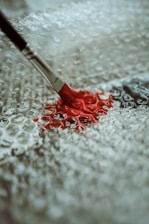 A paintbrush with a metal ferrule and wooden handle is applying red paint onto a textured surface made of bubble wrap, creating an artistic contrast between the smooth paint and bumpy background.