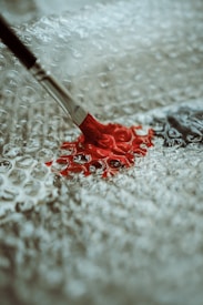 A paintbrush with a metal ferrule and wooden handle is applying red paint onto a textured surface made of bubble wrap, creating an artistic contrast between the smooth paint and bumpy background.
