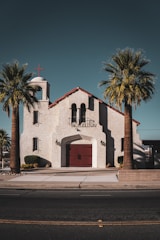 A quaint church with a stucco exterior and red-tiled roof sits in the middle of two palm trees. The building features a bell tower with a red cross at the top. There are large arched windows and a central red double door with metal handles. The pathway leading to the entrance is bordered by small shrubs and neatly trimmed plants.