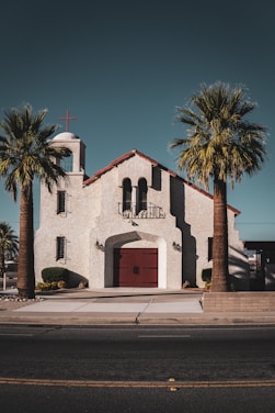 A quaint church with a stucco exterior and red-tiled roof sits in the middle of two palm trees. The building features a bell tower with a red cross at the top. There are large arched windows and a central red double door with metal handles. The pathway leading to the entrance is bordered by small shrubs and neatly trimmed plants.