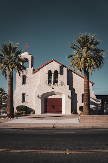 A quaint church with a stucco exterior and red-tiled roof sits in the middle of two palm trees. The building features a bell tower with a red cross at the top. There are large arched windows and a central red double door with metal handles. The pathway leading to the entrance is bordered by small shrubs and neatly trimmed plants.