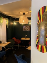 A cozy cafe interior features dark green walls with wooden accents. There are several chairs around small tables, some in navy blue and others in red. The space is illuminated by warm lighting, including a woven pendant lamp. Greenery hangs decoratively from the walls, and a poster of a person is displayed on one wall. A skateboard with a vibrant red and yellow background and an artistic figure is positioned prominently.