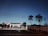 A gas station illuminated in the evening light, with several people attending to vehicles at the pumps. The sky is a deepening blue, and palm trees silhouette against it. Power lines stretch horizontally across the view.
