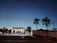 A gas station illuminated in the evening light, with several people attending to vehicles at the pumps. The sky is a deepening blue, and palm trees silhouette against it. Power lines stretch horizontally across the view.