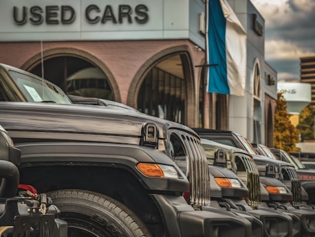 A row of parked black SUVs is prominently displayed in front of a building labeled 'Used Cars.' The vehicles are aligned neatly, showcasing their grills and headlights. The setting suggests a dealership with a modern design, featuring large arched windows and a building facade with neutral tones. A flagpole with a blue and white flag is visible, adding a touch of color to the otherwise monochromatic scene. The sky is overcast, with clouds creating a diffused light that softens the overall atmosphere.