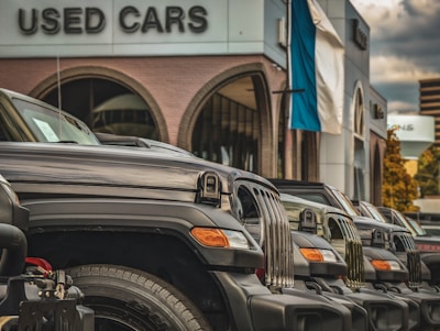 A row of parked black SUVs is prominently displayed in front of a building labeled 'Used Cars.' The vehicles are aligned neatly, showcasing their grills and headlights. The setting suggests a dealership with a modern design, featuring large arched windows and a building facade with neutral tones. A flagpole with a blue and white flag is visible, adding a touch of color to the otherwise monochromatic scene. The sky is overcast, with clouds creating a diffused light that softens the overall atmosphere.