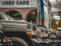 A row of parked black SUVs is prominently displayed in front of a building labeled 'Used Cars.' The vehicles are aligned neatly, showcasing their grills and headlights. The setting suggests a dealership with a modern design, featuring large arched windows and a building facade with neutral tones. A flagpole with a blue and white flag is visible, adding a touch of color to the otherwise monochromatic scene. The sky is overcast, with clouds creating a diffused light that softens the overall atmosphere.