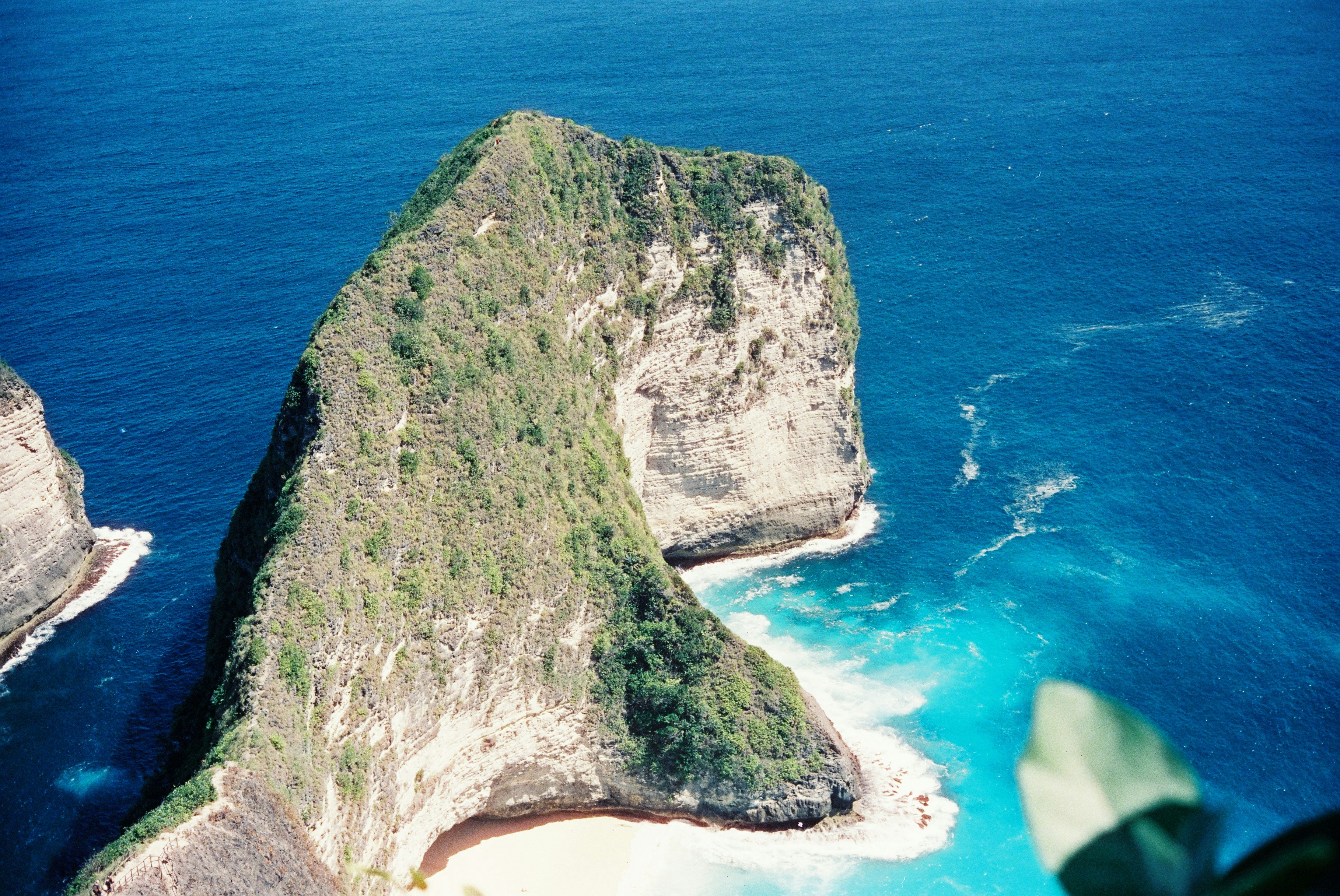 Aerial photograph of a green-clad rock island jutting into bright turquoise waters, with white surf along the shore.