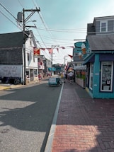 A quaint street lined with shops featuring outdoor signage, including a large ice cream cone. American flags and a Budweiser sign add to the small-town atmosphere. The street is busy with pedestrians and a bicycle cart labeled 'bwell' in the center.