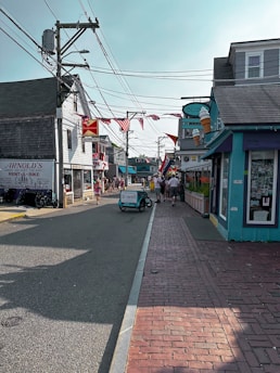 A quaint street lined with shops featuring outdoor signage, including a large ice cream cone. American flags and a Budweiser sign add to the small-town atmosphere. The street is busy with pedestrians and a bicycle cart labeled 'bwell' in the center.