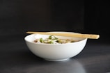 A white ceramic bowl filled with a noodle dish, garnished with green herbs and possibly onions. A pair of wooden chopsticks rests across the top of the bowl. The background is a dark color that contrasts with the simplicity and elegance of the bowl.