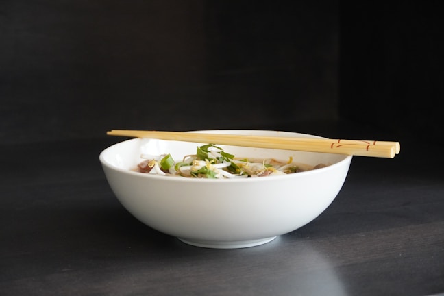 A white ceramic bowl filled with a noodle dish, garnished with green herbs and possibly onions. A pair of wooden chopsticks rests across the top of the bowl. The background is a dark color that contrasts with the simplicity and elegance of the bowl.