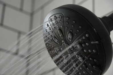 A black showerhead with multiple tiny openings is mounted against a tiled wall. Water streams are visibly spraying out in several directions, capturing a dynamic aspect of flowing water.