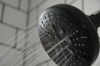 A black showerhead with multiple tiny openings is mounted against a tiled wall. Water streams are visibly spraying out in several directions, capturing a dynamic aspect of flowing water.